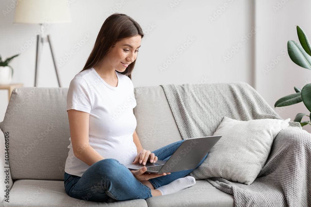 Smiling pregnant millennial woman relaxing with laptop at home, browsing internet