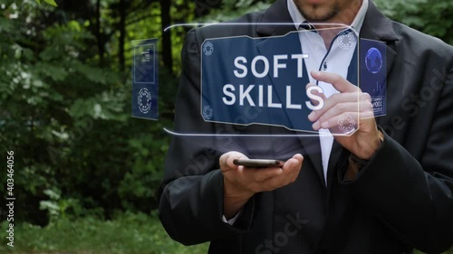 Unrecognizable businessman activates conceptual HUD holograms on smartphone with text Soft Skills. Bearded man in white shirt and acket with holographic screen on a background of green trees photo