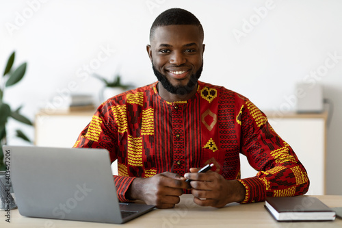 African Freelancer Guy In Traditional Costume Sitting At Desk At Home Office photo