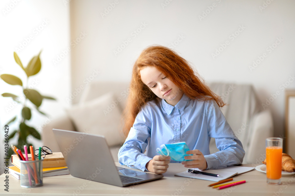 Sad girl holding medical mask studying at home with laptop Sad girl holding medical mask studying at home with laptop