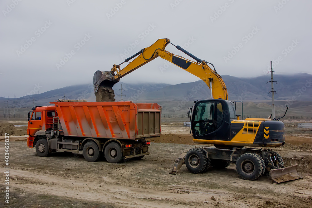 Excavator at a construction site loads a truck against a background of mountains Excavator at a construction site loads a truck against a background of mountains