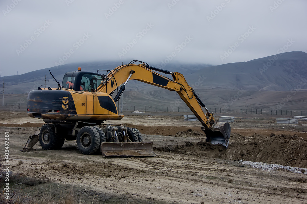 Yellow excavator digs a trench against the backdrop of mountains. Yellow excavator digs a trench against the backdrop of mountains.