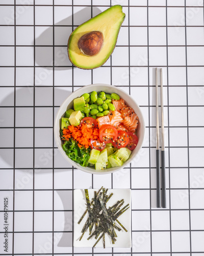 Poke bowl with salmon fish, avocado and other vegetables with chopsticks top view on geometric background.