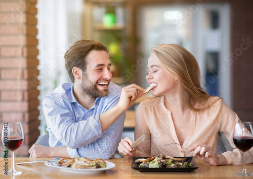 Joyful millennial guy feeding delicious salad to his beloved woman during festive dinner at cafe