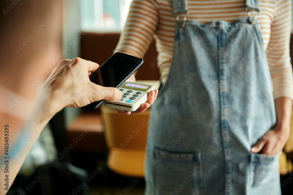 Woman wearing face mask behind cafe counter holding contactless payment device while customer uses mobile phone to pay bill Woman wearing face mask behind cafe counter holding contactless payment device while customer uses mobile phone to pay bill
