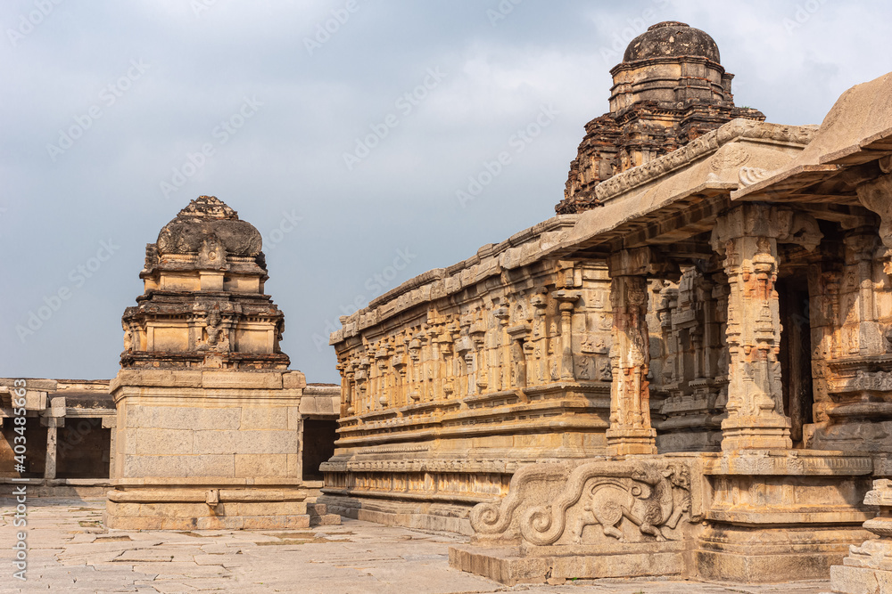 Hampi, Karnataka, India - November 5, 2013: Sri Krishna temple in ruins. Yellow-brown shrine on side of facade with steps to enter, sculpted pillars, and dome vimanam under light blue cloudscape.