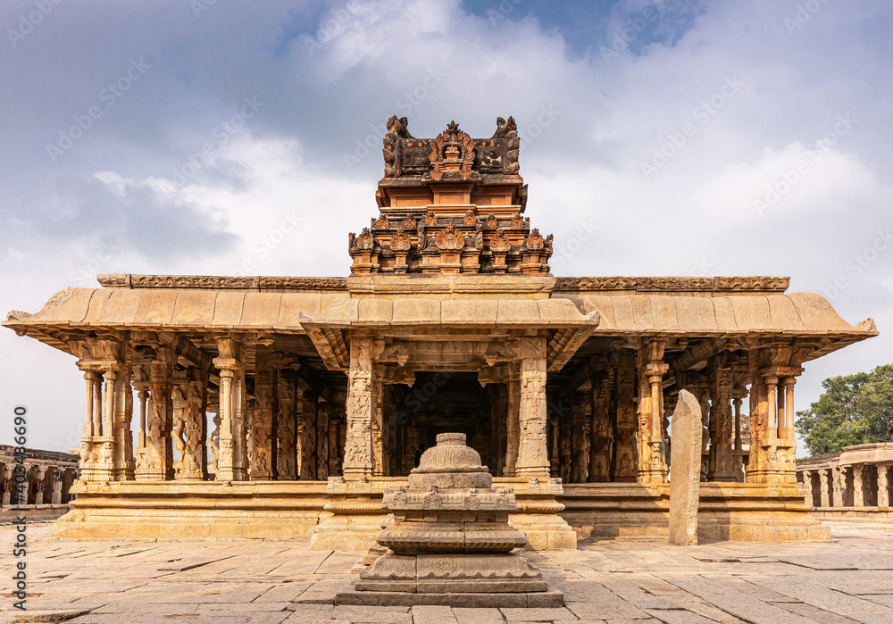Obraz premium Hampi, Karnataka, India - November 5, 2013: Sri Krishna temple in ruins. Red stone vimanam on top of yellow stone mandapam in front of sanctum under blue cloudscape with some green foliage.