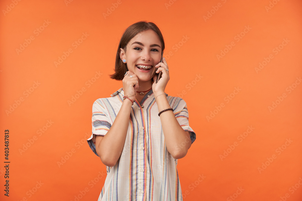 Female student talk on smart phone, wearing striped shirt, teeth braces and bracelets, hear a good news. Smiling girl looking happy and watching at the camera isolated over orange background Female student talk on smart phone, wearing striped shirt, teeth braces and bracelets, hear a good news. Smiling girl looking happy and watching at the camera isolated over orange background