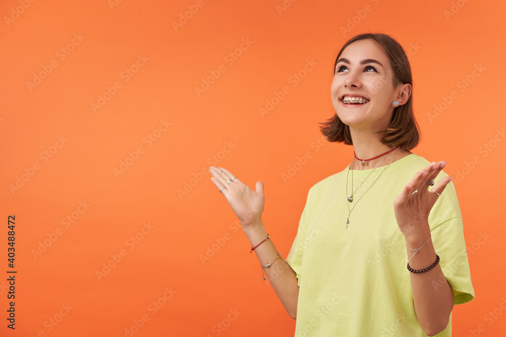 Teenage girl, cheerful and happy looking woman with brunette short hair. Happy to share feelings, feels lucky. Watch to the left and up at the copy space over orange background. Wearing green t-shirt Teenage girl, cheerful and happy looking woman with brunette short hair. Happy to share feelings, feels lucky. Watch to the left and up at the copy space over orange background. Wearing green t-shirt