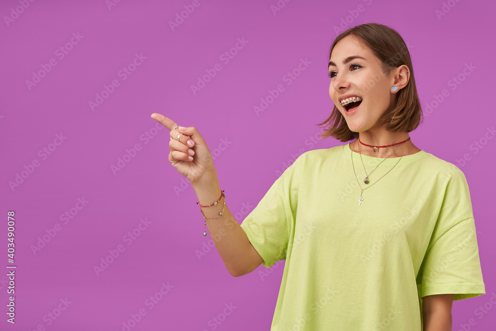 Female student, young lady, laughing and pointing her finger to the left at the copy space over purple background. Showing a sign. Wearing green t-shirt, teeth braces, bracelets and rings Female student, young lady, laughing and pointing her finger to the left at the copy space over purple background. Showing a sign. Wearing green t-shirt, teeth braces, bracelets and rings