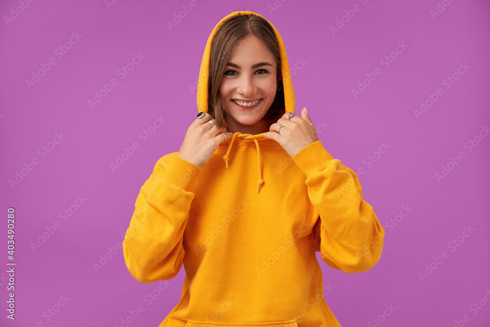 Portrait of attractive, nice looking girl with short brunette hair. Smiling at he camera and touching hood with hands, over purple background. Wearing orange hoodie, rings and teeth braces Portrait of attractive, nice looking girl with short brunette hair. Smiling at he camera and touching hood with hands, over purple background. Wearing orange hoodie, rings and teeth braces