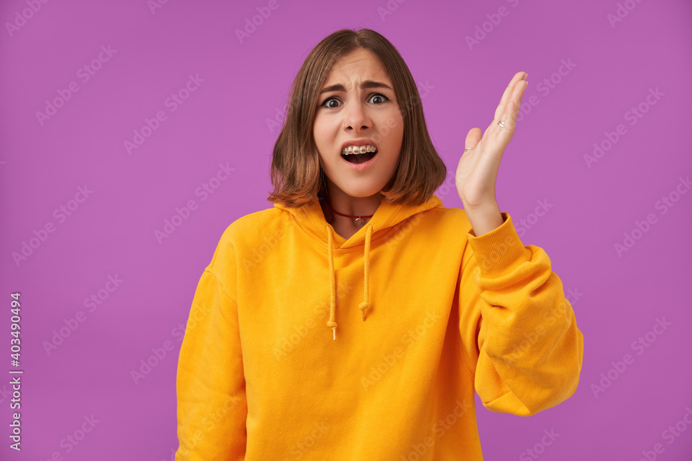 Portrait of attractive, nice looking girl with short brunette hair, shocked, horrified lady lifts her hand. Looking at the camera over purple background. Wearing orange hoodie, teeth braces and rings Portrait of attractive, nice looking girl with short brunette hair, shocked, horrified lady lifts her hand. Looking at the camera over purple background. Wearing orange hoodie, teeth braces and rings