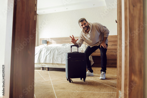 Smiling bearded man in sweatshirt standing in hotel room and showing victory sign. He is on a weekend. photo