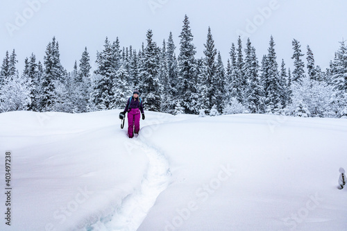 Woman walking, hiking in deep snowy woods during winter time surrounded by white covered snowy trees and wearing pink pants, purple jacket standing out from the whiteness.