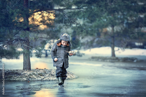 Cute funny little boy in cap with ear-flaps playing with toy little ship and puddles in yearly spring during the sunset in Russia. Image with toning and selective focus