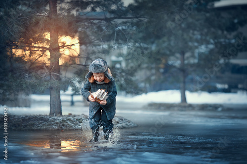 Fototapeta Naklejka Na Ścianę i Meble -  Cute funny little boy in cap with ear-flaps playing with toy little ship and puddles in yearly spring during the sunset in Russia. Image with toning and selective focus
