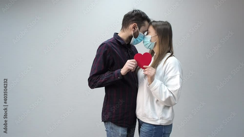 A young couple in protective medical masks touched their heads, red paper heart they hold in their hands. A young couple in protective medical masks touched their heads, red paper heart they hold in their hands.