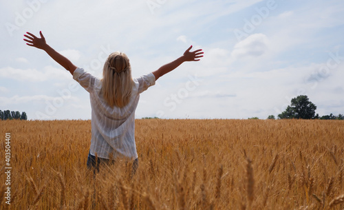 A young woman in a white shirt enjoys the moment among the golden wheat field against the blue sky. View from the back. Place for text.