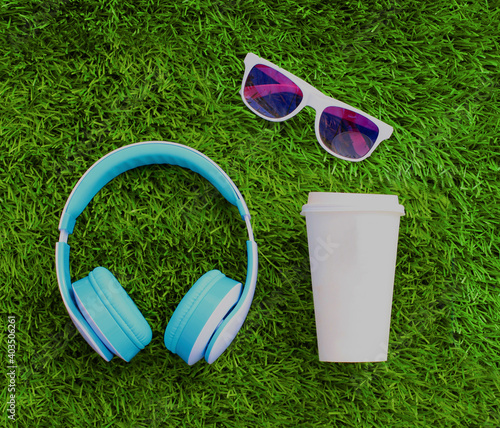 Blue wireless headphones, coffee cup, sunglasses on a green grass background, top view
