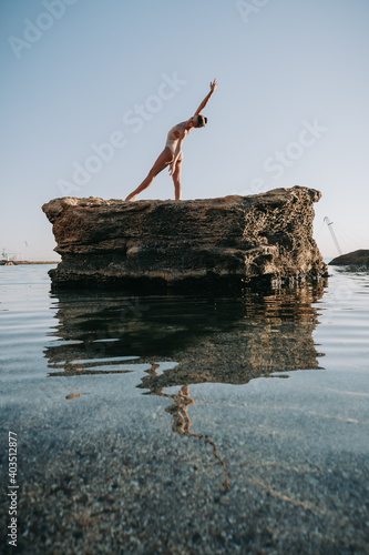 Young ballerina in beige swimsuit dancing ballet on sea or ocean sandy beach in morning light. Concept of stretching, art, nature beauty.