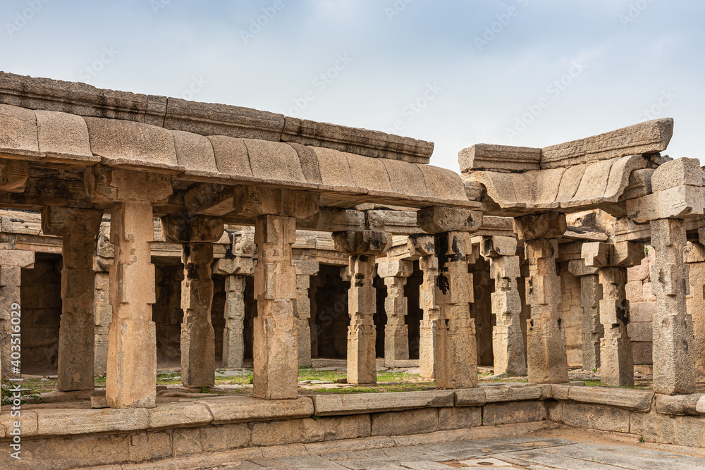 Hampi, Karnataka, India - November 5, 2013: Sri Krishna temple in ruins. Beige stone pillars and beams at a ruinous corner of the premises under light blue sky. Some grass.