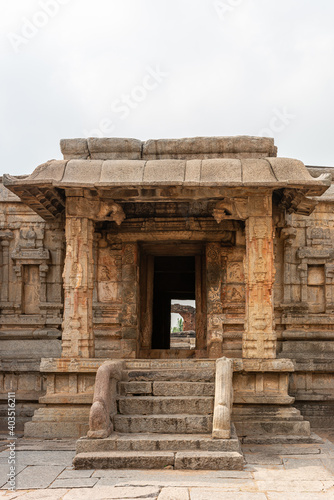 Hampi, Karnataka, India - November 5, 2013: Sri Krishna temple in ruins. Brown stone steps with balusters lead to entrance to inner sanctum under silver sky. Look through shows green foliage.