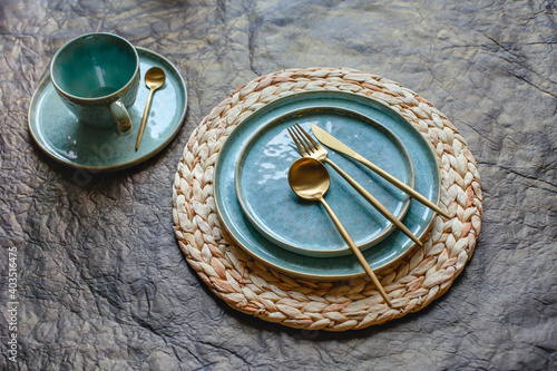 Composition from a turquoise plate and cup, golden cutlery and wicker plate stand on brown background, top view, horizontal image. © Hanna