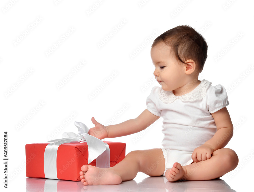 Baby and wrapped festive gift box with bow. Cute interested curious infant girl child in jumpsuit sitting on white floor studio background looking at present. Birthday or Christmas celebration concept Baby and wrapped festive gift box with bow. Cute interested curious infant girl child in jumpsuit sitting on white floor studio background looking at present. Birthday or Christmas celebration concept