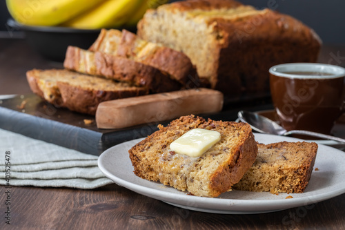 Close up of slices of banana bread with butter on a small plate with a sliced banana bread loaf in behind.