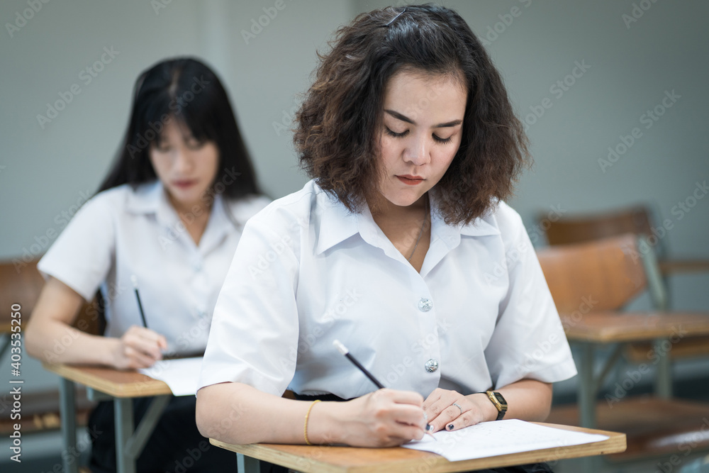 Selective focus of the teenage college students sit on lecture chairs do final examination and write on examination paper answer sheets in the classroom. University students in uniform in classroom. Selective focus of the teenage college students sit on lecture chairs do final examination and write on examination paper answer sheets in the classroom. University students in uniform in classroom.