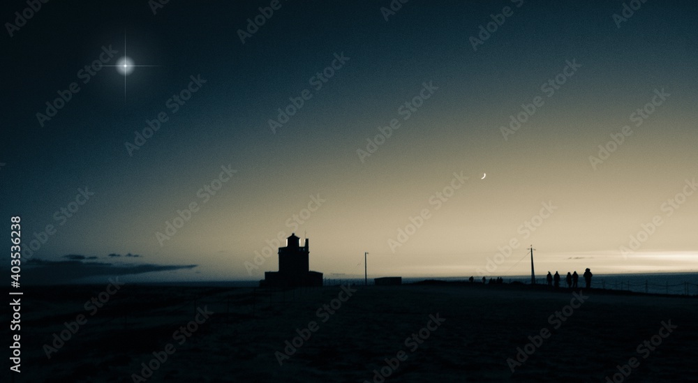 Greyscale photo of a lighthouse overlooking the ocean on a winter's night. With a crescent moon and aeed bright star.