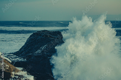 A large wave crashing against a rock on the Icelandic coast, with the ocean on the horizon. During dusk on a cold winter day.