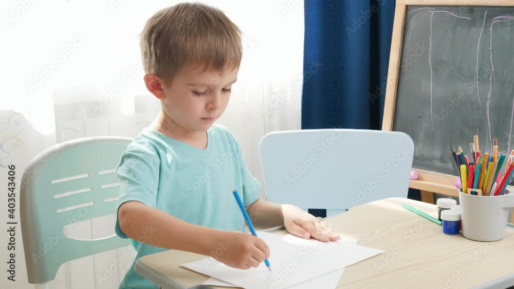 Concentrated little boy drawing or writing with pencil while sitting behind school desk at home or school classroom. Concept of education and remote school at home during lockdown and self isolation Concentrated little boy drawing or writing with pencil while sitting behind school desk at home or school classroom. Concept of education and remote school at home during lockdown and self isolation