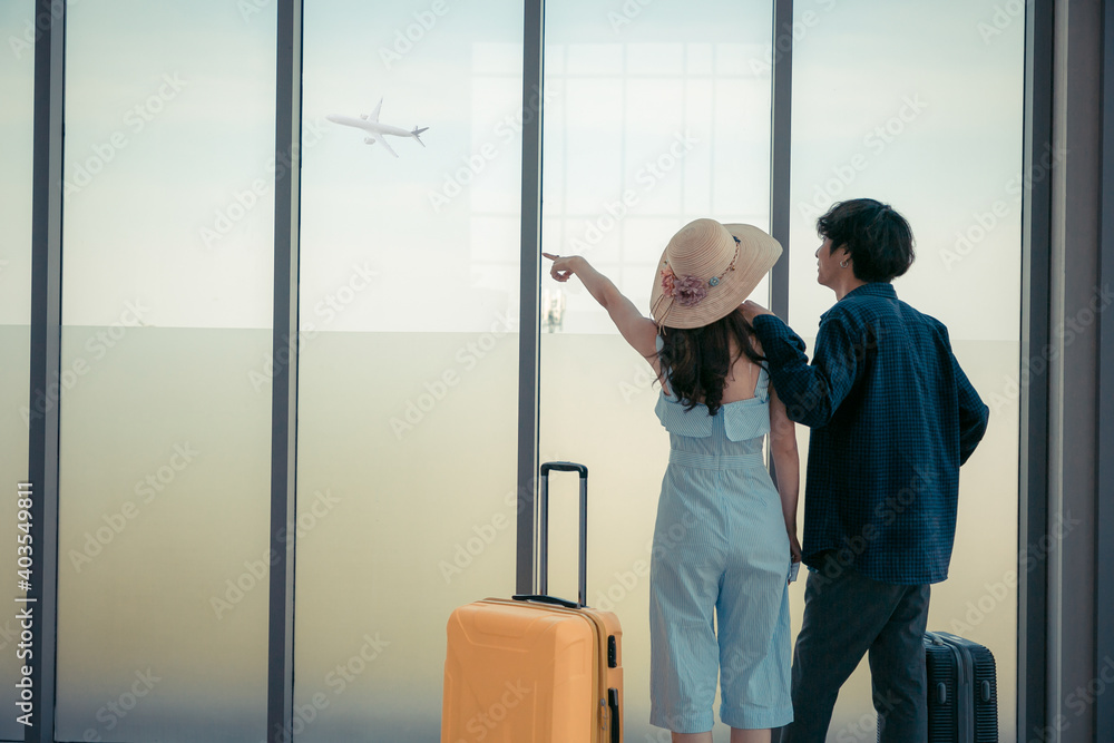 Couple love is looking at flying plain in sky. They are standing near window at airport and holding hands.