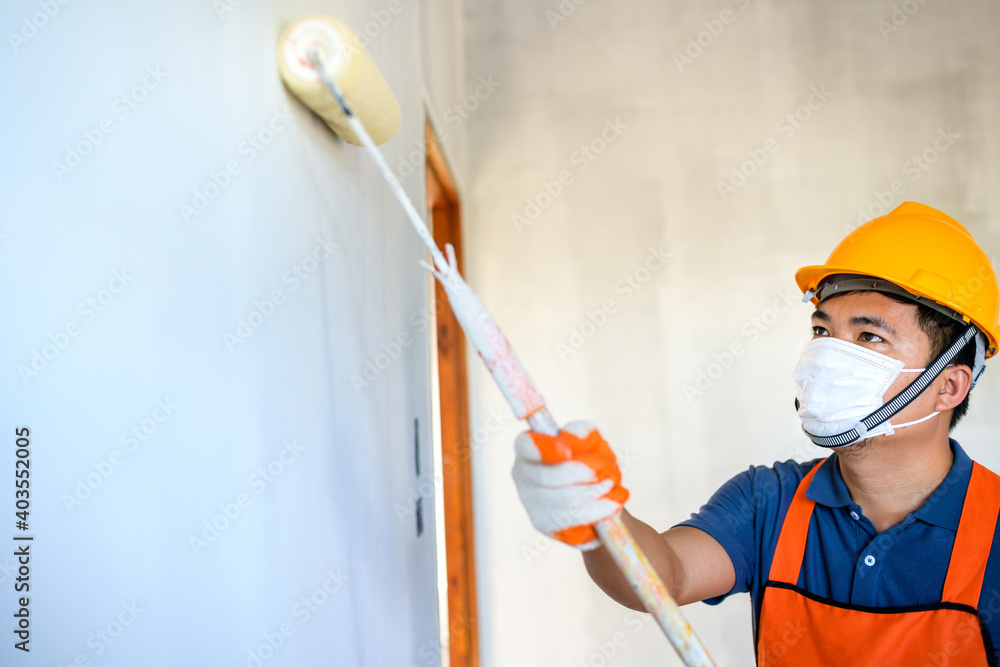 Young Asian painter Wear a protective mask to paint the indoor white walls with a paint roller in the new house.