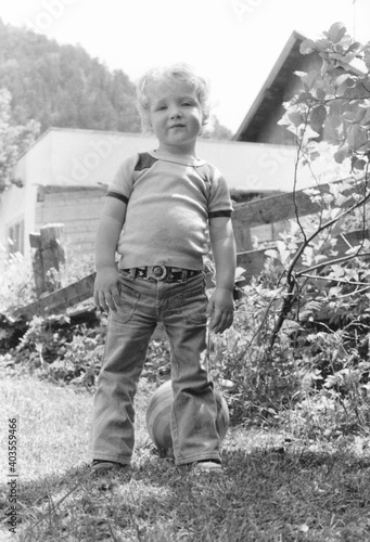 Vintage monochrome 1978 low angle image of a young boy with ball wearing flare pant jeans and tshirt looking at camera