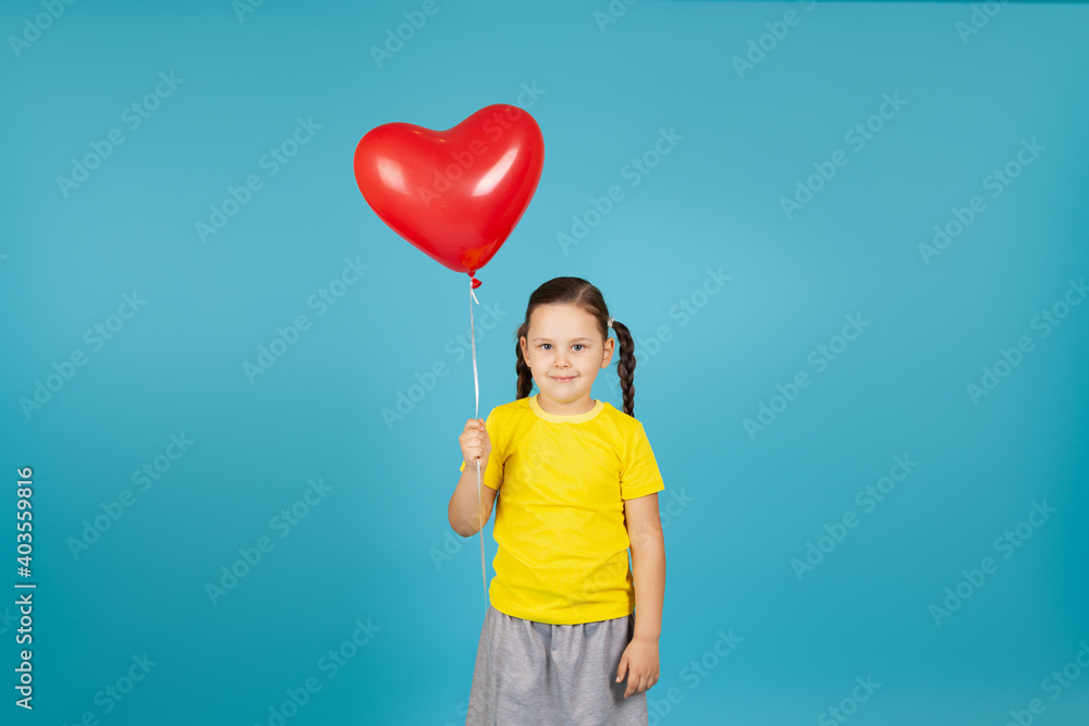 cute, demure girl with pigtails in a yellow T-shirt holds a flying red balloon in the shape of a heart , isolated on a blue background. cute, demure girl with pigtails in a yellow T-shirt holds a flying red balloon in the shape of a heart , isolated on a blue background.