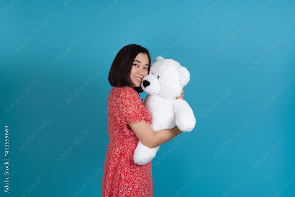 mock up Asian woman in red dress sideways to camera gently cuddles white Teddy bear isolated on blue background. mock up Asian woman in red dress sideways to camera gently cuddles white Teddy bear isolated on blue background.