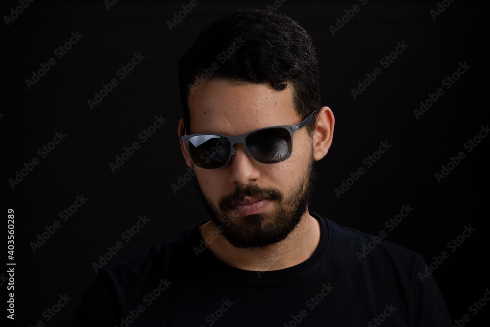 Portrait of a young man in a studio shot. He is caucasian with casual beard and is serious looking to one side, he si wearing sunglasses. Portrait of a young man in a studio shot. He is caucasian with casual beard and is serious looking to one side, he si wearing sunglasses.