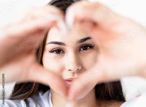 Young happy brunette woman in white t-shirt showing heart sign with her hands in front of face