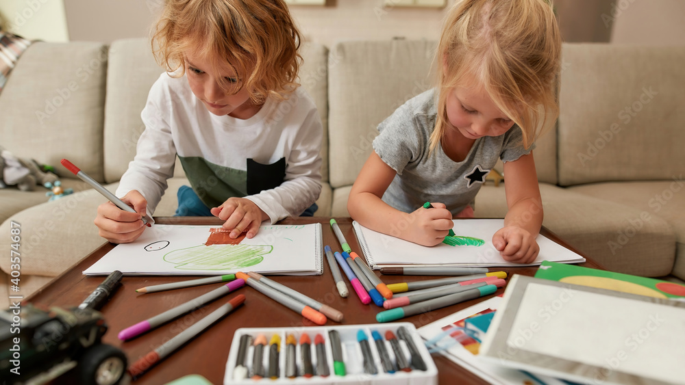 Focused little siblings, boy and girl drawing on paper using marker pen while spending time together at home Focused little siblings, boy and girl drawing on paper using marker pen while spending time together at home