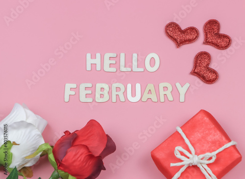 flat lay of wooden letters HELLO FEBRUARY  with  red and white roses, red glitter hearts and red gift box on pink background.