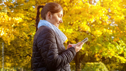 young woman in a black jacket and a gray scarf is typing a message on the phone against the background of yellowed trees copyspace