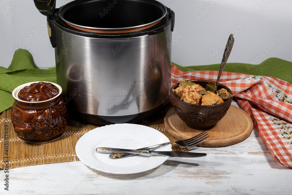 On the table is a slow cooker, a bowl with a spoon and cooked meat, a ceramic pot, and a plate with a knife and fork. On the table is a slow cooker, a bowl with a spoon and cooked meat, a ceramic pot, and a plate with a knife and fork.