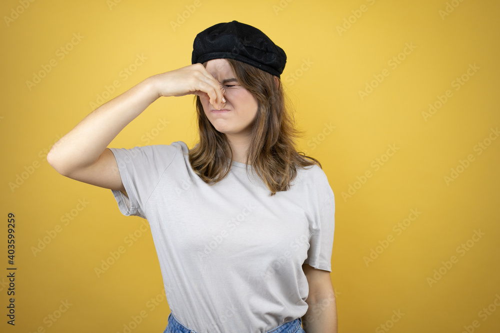 Beautiful young caucasian girl wearing french look with beret over isolated yellow background smelling something stinky and disgusting, intolerable smell, holding breath with fingers on nose Beautiful young caucasian girl wearing french look with beret over isolated yellow background smelling something stinky and disgusting, intolerable smell, holding breath with fingers on nose