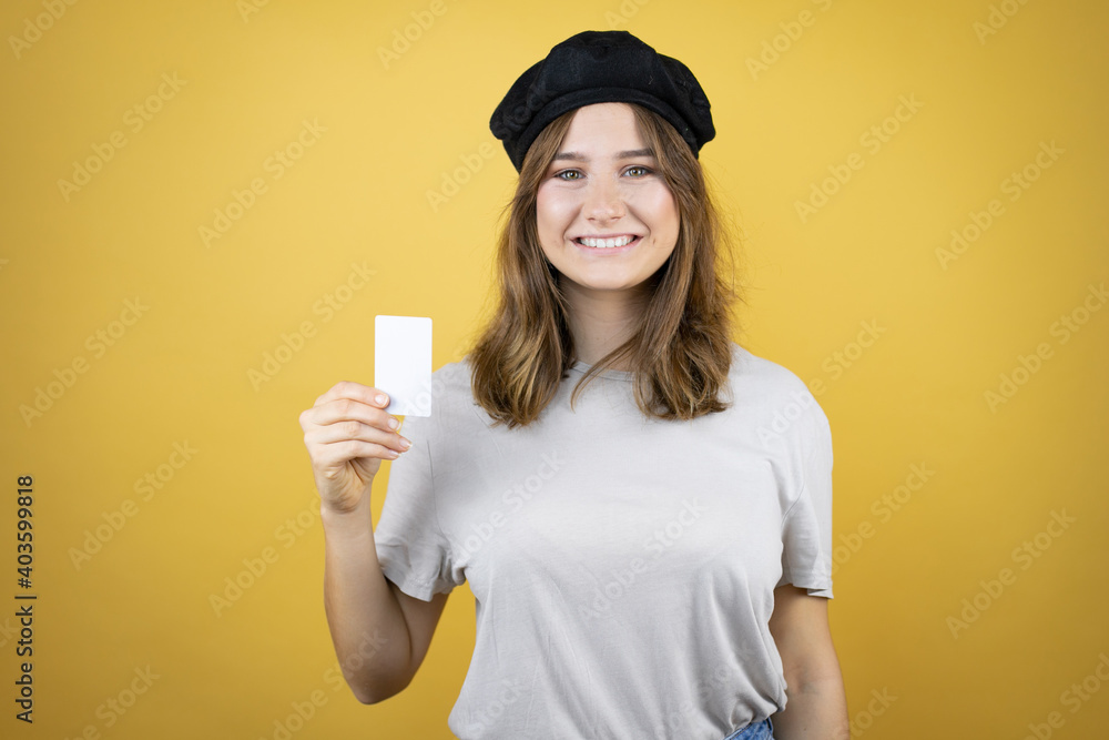 Beautiful young caucasian girl wearing french look with beret over isolated yellow background smiling and holding white card Beautiful young caucasian girl wearing french look with beret over isolated yellow background smiling and holding white card