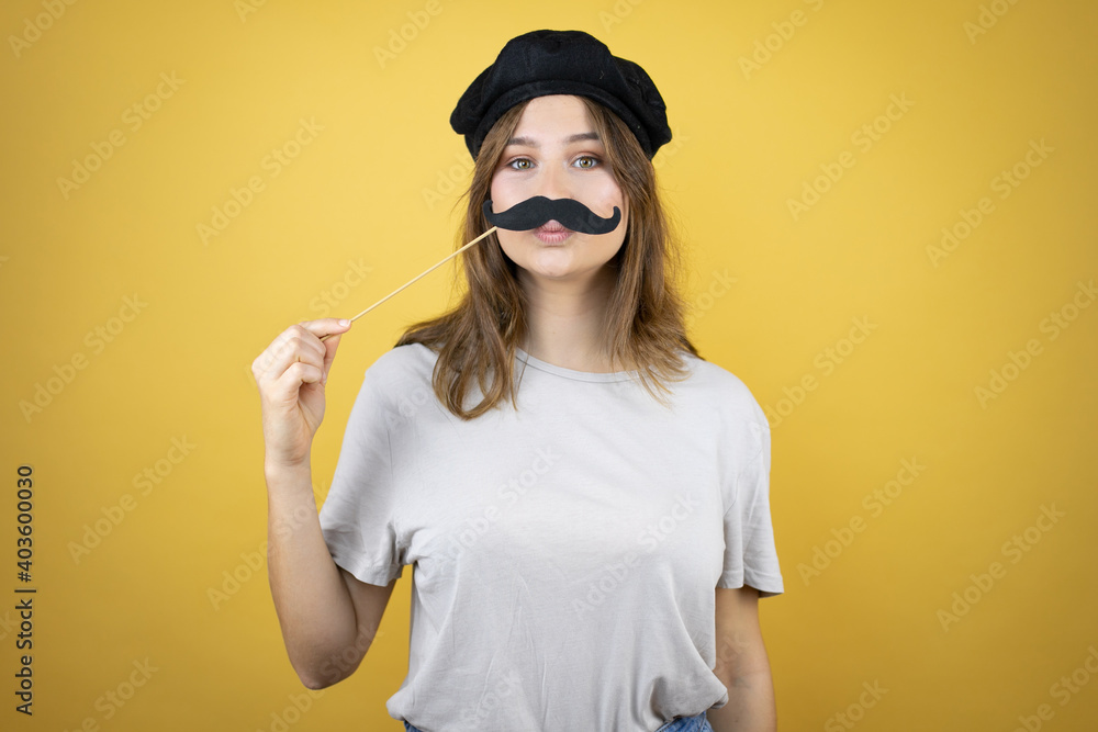 Beautiful young caucasian girl wearing french look with beret over isolated yellow background happy with mustache mask Beautiful young caucasian girl wearing french look with beret over isolated yellow background happy with mustache mask