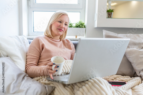 Beautiful young woman drinking coffee at home in her bed wearing a cozy t shirt while working on her laptop. Checking email in the morning, freelance concept