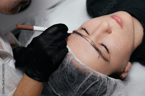 Markup with pencil on eyebrows of young girl while permanent make up. Eyebrow permanent makeup procedure  microblading. Beauty Concept. Eyebrow correction  threading. Cosmetologist