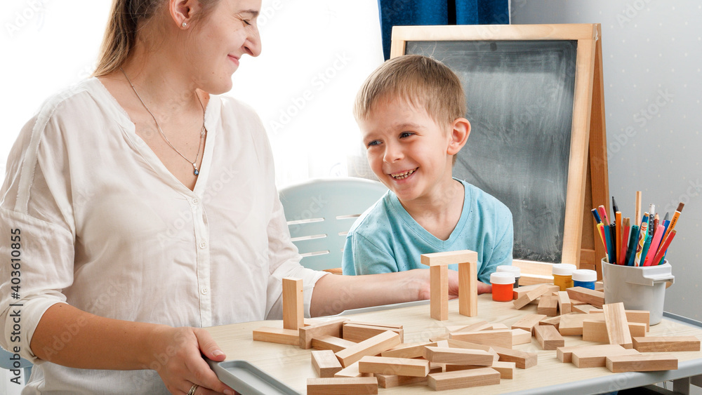 Happy laughing boy playing in wooden blocks and bricks with young mother